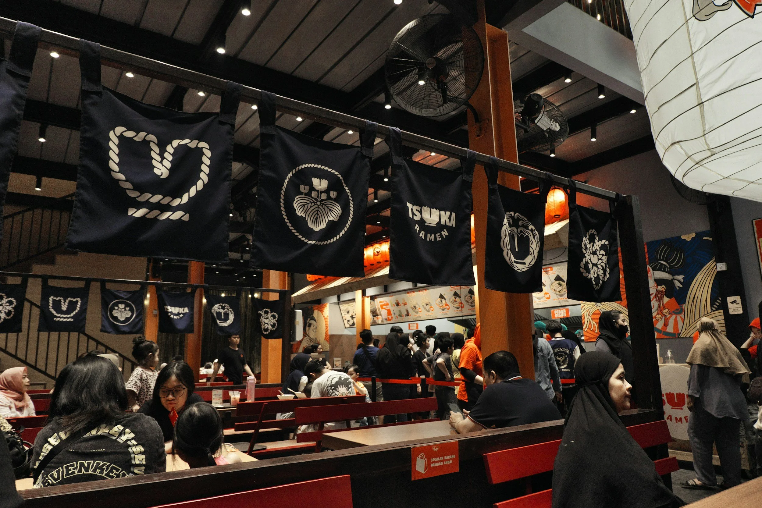 Inside a busy Asian restaurant with many customers, black banners with white symbols and text, and a menu on the wall. The ceiling has black beams, fans, and warm lighting.