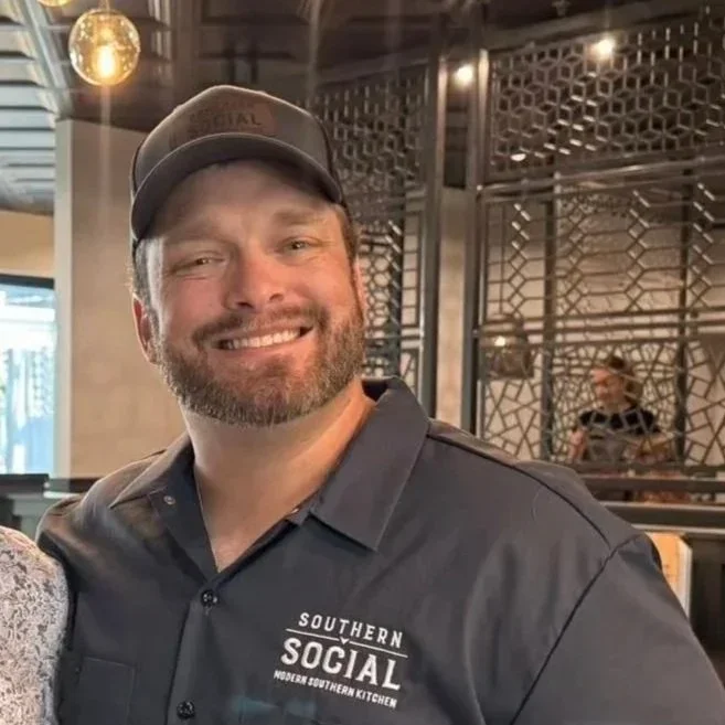 A smiling man wearing a gray cap and a dark gray shirt with the logo 'Southern Social Modern Southern Kitchen' inside a restaurant.