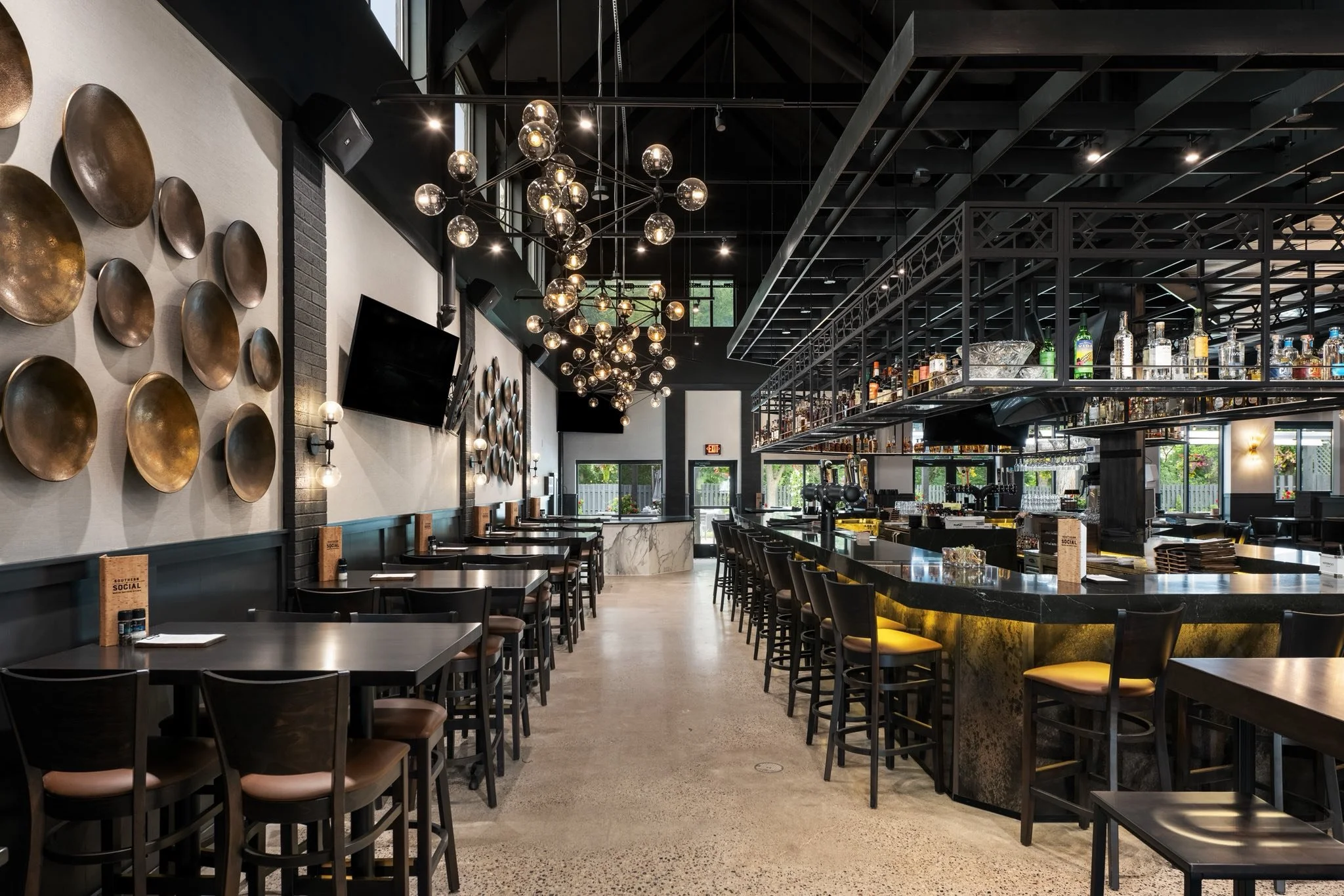 Interior of a modern bar or restaurant with black walls, gold wall art, multiple spherical light fixtures hanging from the ceiling, a marble-topped bar with liquor bottles, and a row of chairs along the bar and tables with menus.