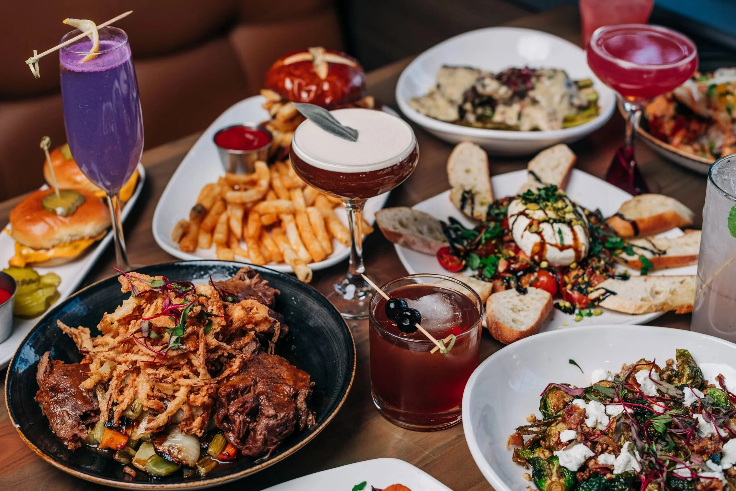 Assorted dishes and drinks on a table, including pulled pork with fried onions, French fries, various salads, and several colorful cocktails.