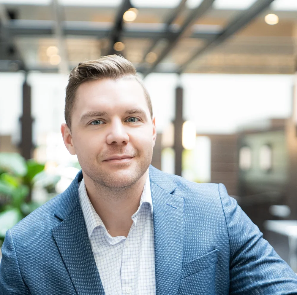 Headshot of a young man with light skin, short brown hair, blue eyes, wearing a blue blazer and checkered shirt, in a modern indoor setting with natural light.