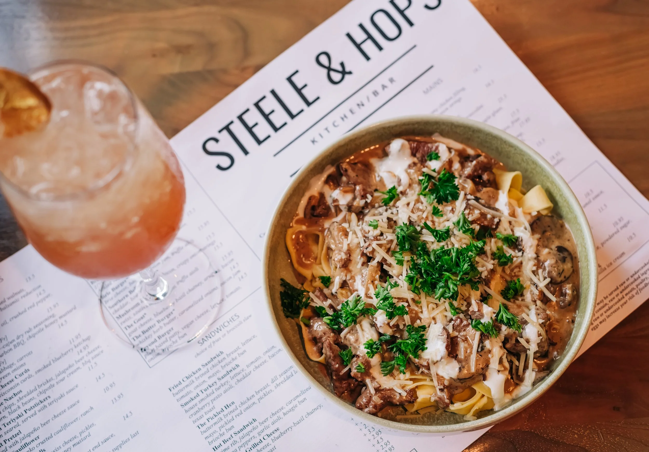 A bowl of cheesy, meaty pasta garnished with parsley, sitting on a menu and a wooden table, with a drink glass nearby.