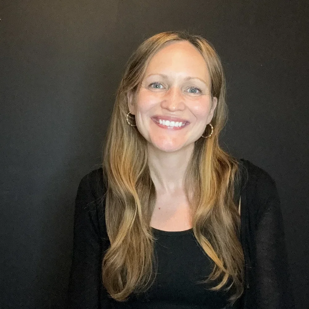 A woman with long, wavy blonde hair, blue eyes, and hoop earrings smiling in front of a dark background.