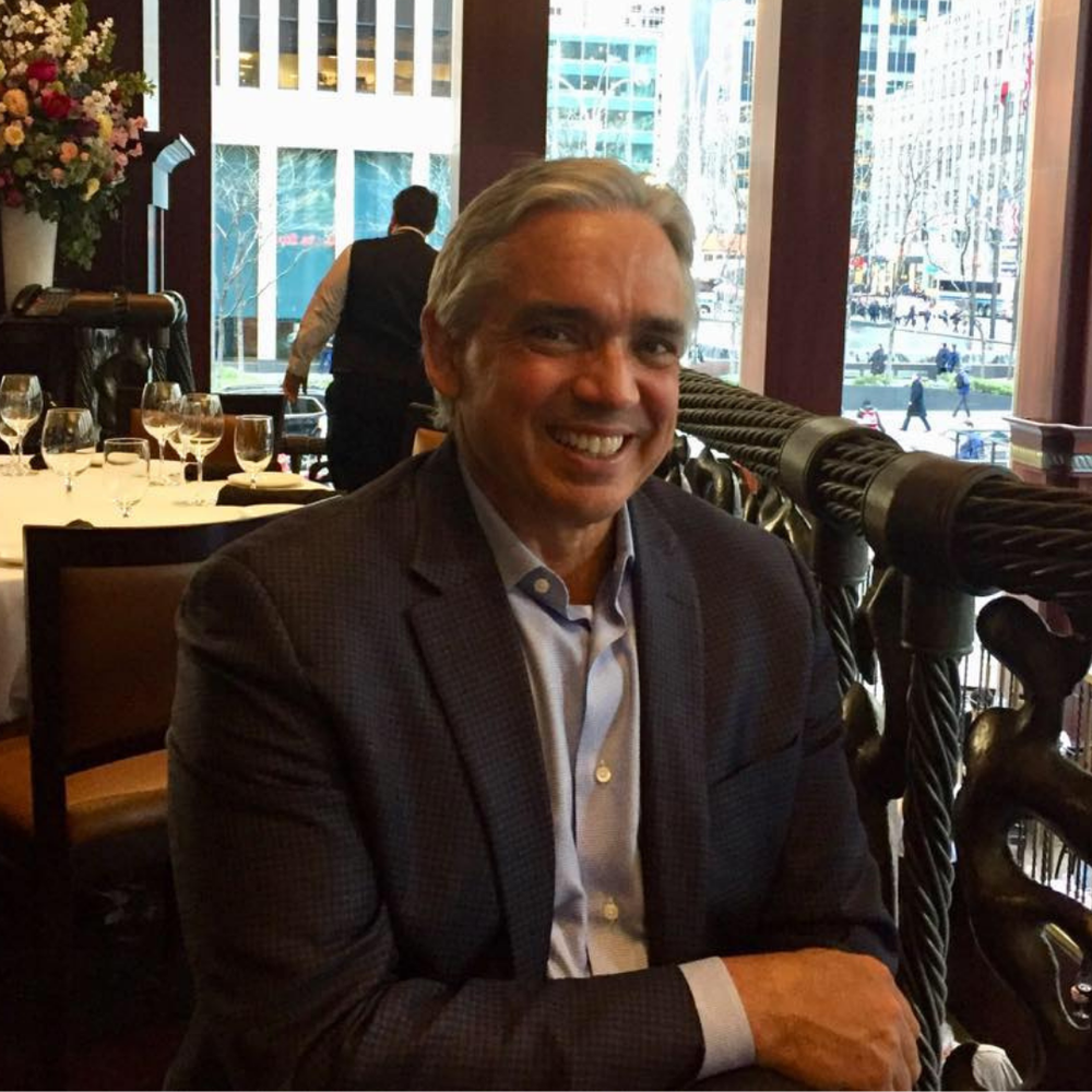 A smiling man in a suit sitting at a table in a restaurant with a city street view outside the window.