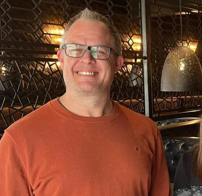 A smiling man with glasses and light hair, wearing an orange shirt, in a restaurant with modern interior decor, including pendant lights and a decorative partition in the background.