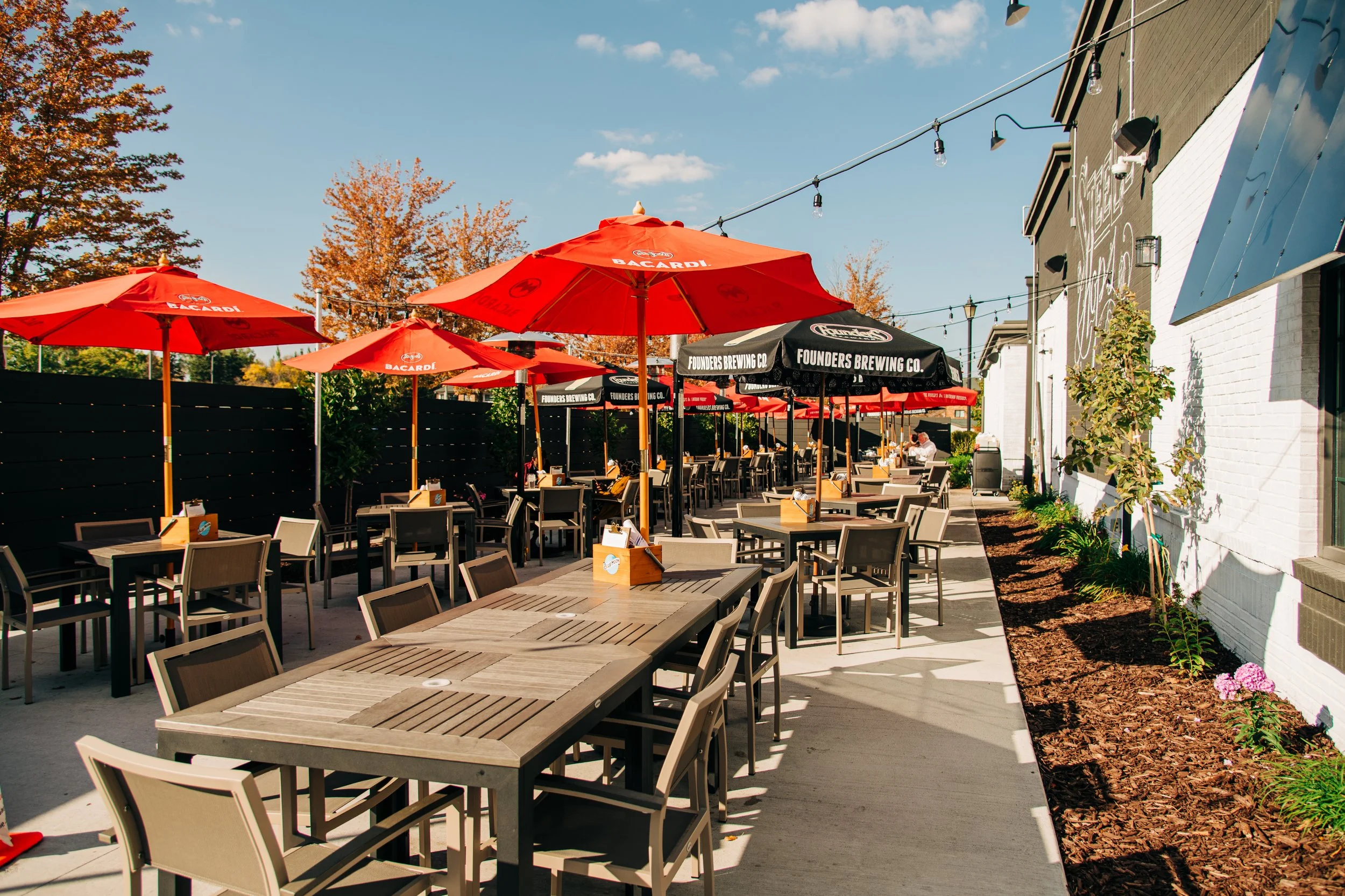 Outdoor patio seating area with several wooden tables and beige chairs, red umbrellas with Bacardi logos, string lights, a black fence, a white brick building with a mural, and a tree-lined sidewalk under a blue sky.