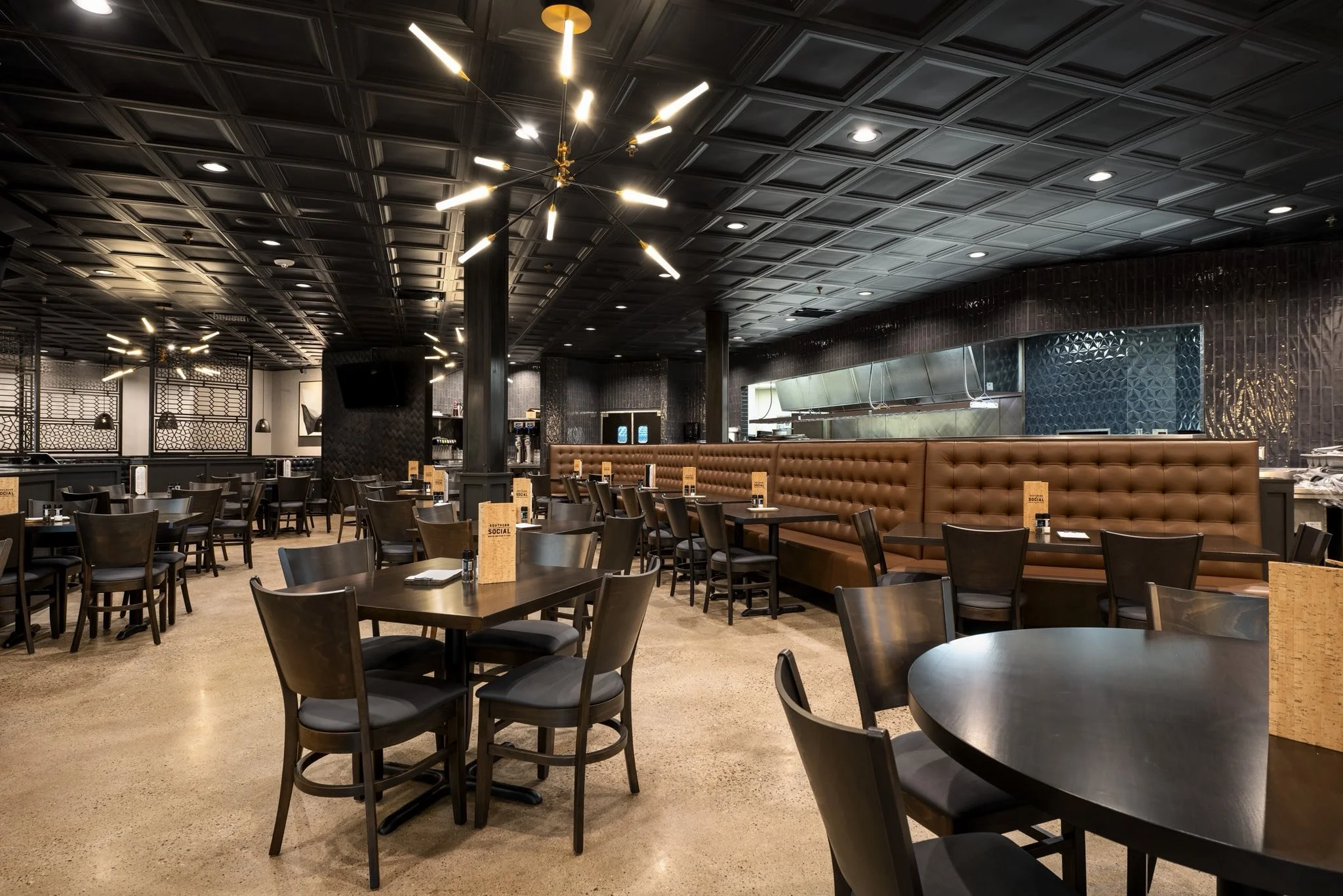 An empty restaurant dining area with black tables and chairs, brown upholstered booth seating, dark ceiling with modern lighting fixtures, and decorative wall panels.
