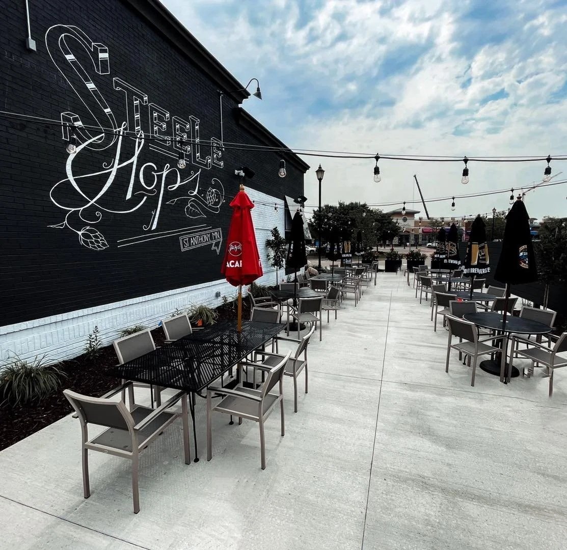 Empty outdoor patio of a restaurant with tables, chairs, umbrellas, and string lights, against a brick building with a large decorative Sign that reads 'Stelle Hop's'.