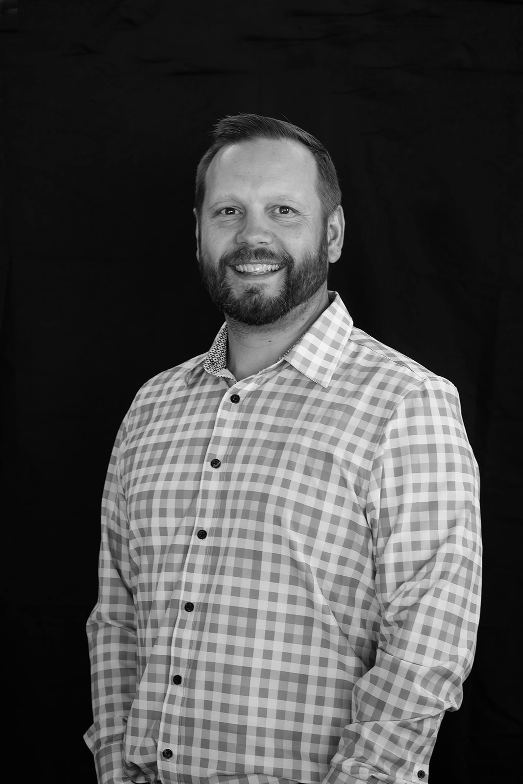 Black and white portrait of a man smiling, wearing a checkered shirt, with a dark background.