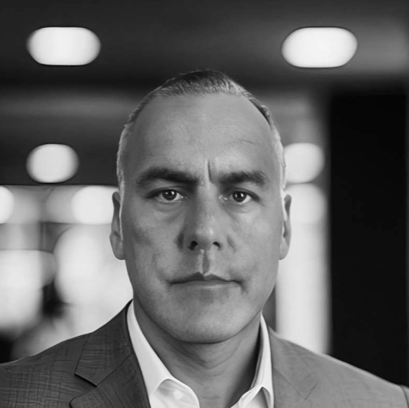 Black and white photo of a man in a suit with a serious expression, indoors with blurred lights in the background.