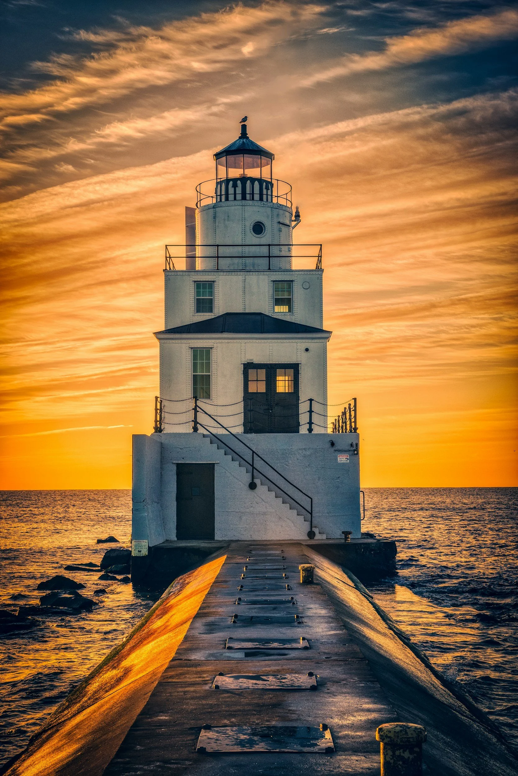 A lighthouse on a pier at sunset with a dramatic sky and ocean in the background.