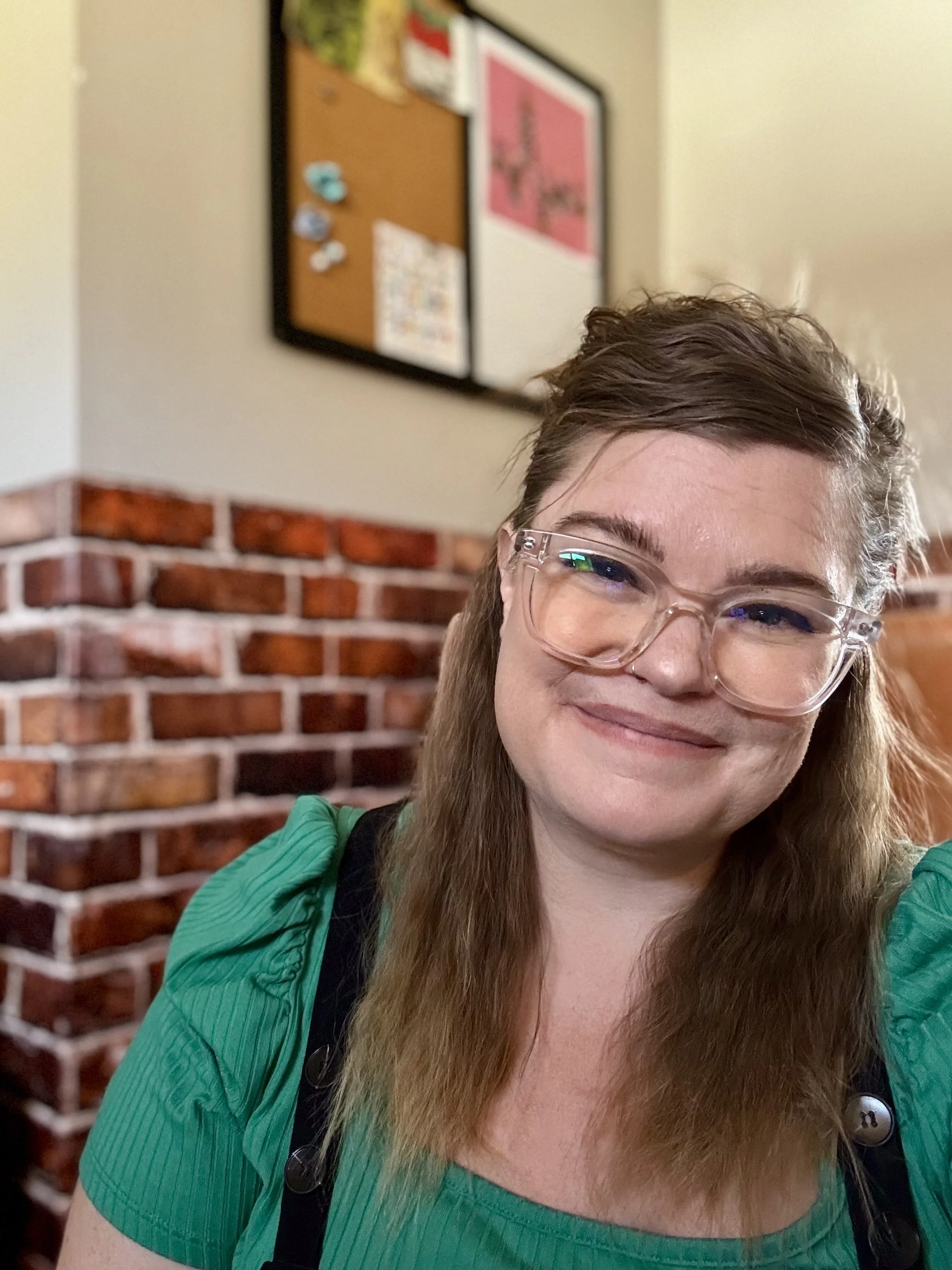 A smiling woman with brown hair and wearing clear glasses, a green shirt, and a black strap across her shoulder, sitting inside a room with a brick wall and bulletin board in the background.