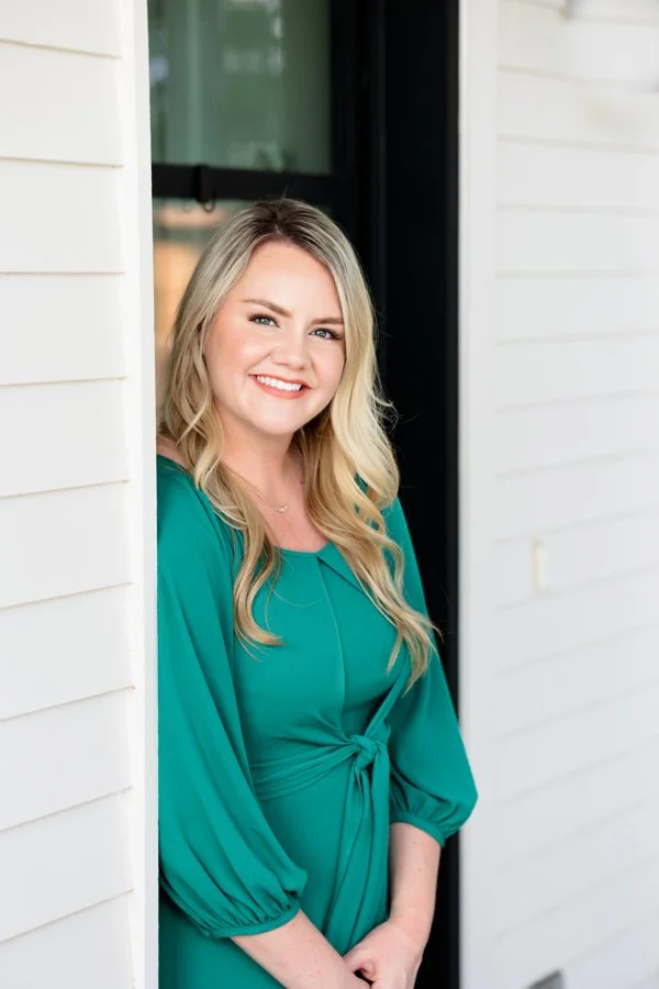 A woman with long blonde hair smiling, standing at a white siding wall near a door in a teal dress with puffy sleeves.