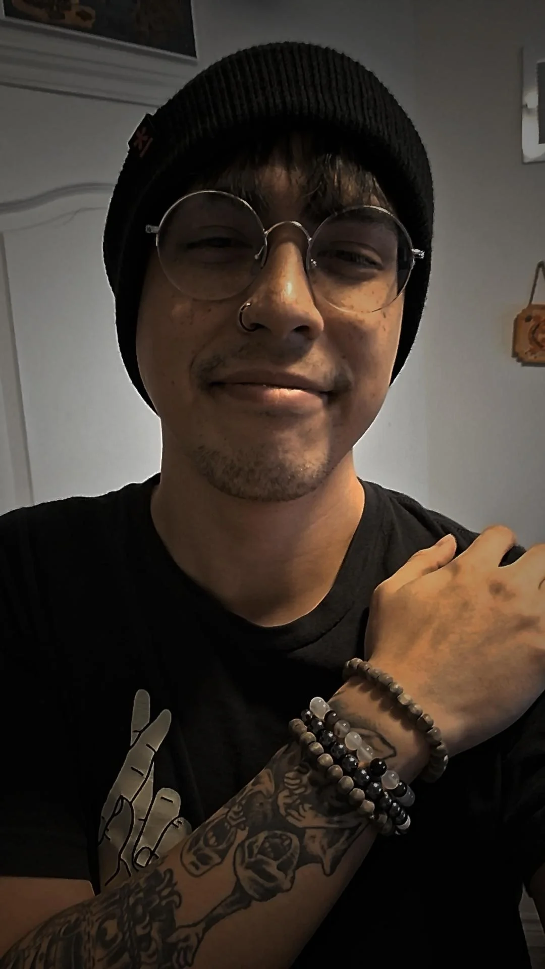 A young man with glasses, a nose ring, and tattoos on his arm, wearing a black beanie and black T-shirt, smiling with his hand resting on his shoulder.