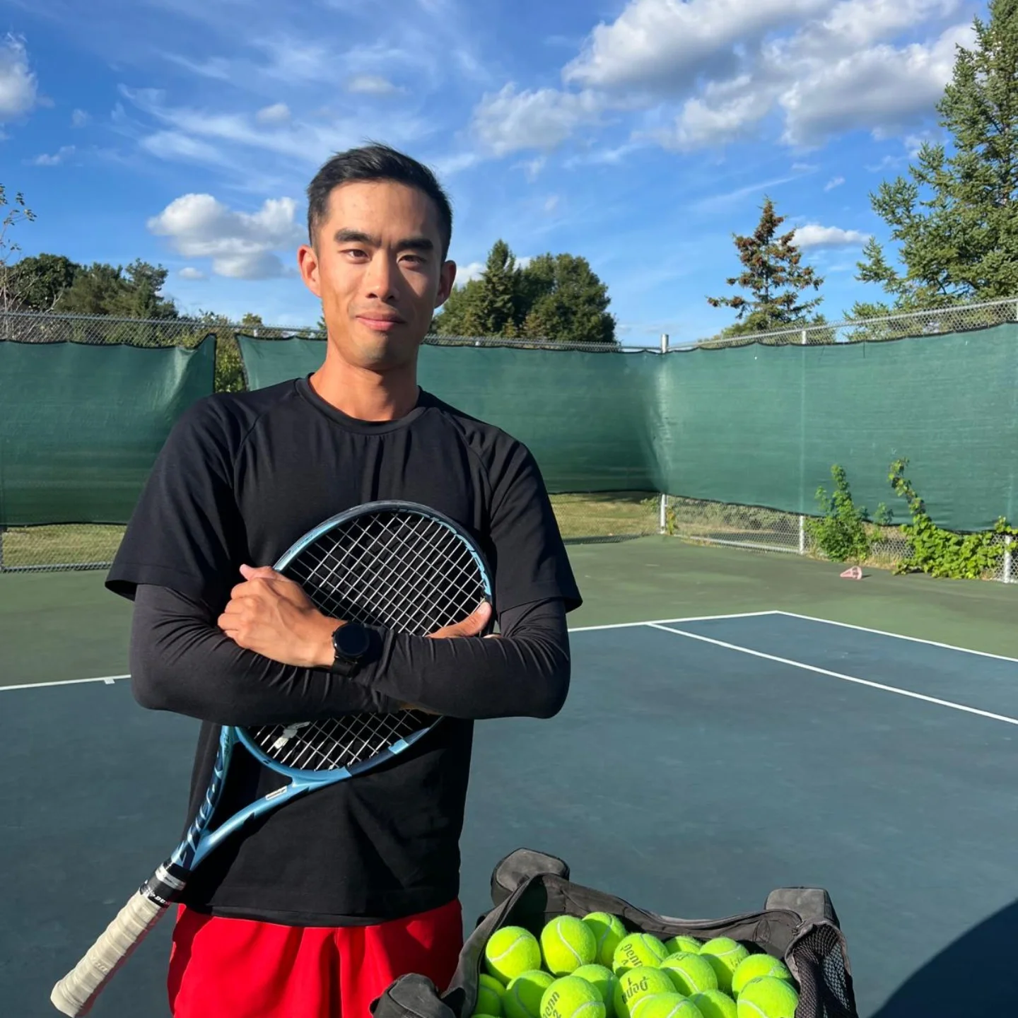 Coach Wesley Tsang smiling on a tennis court holding a tennis racquets, standing next to a bag of tennis balls, with a green windscreen and trees in the background under bright sunny sky.