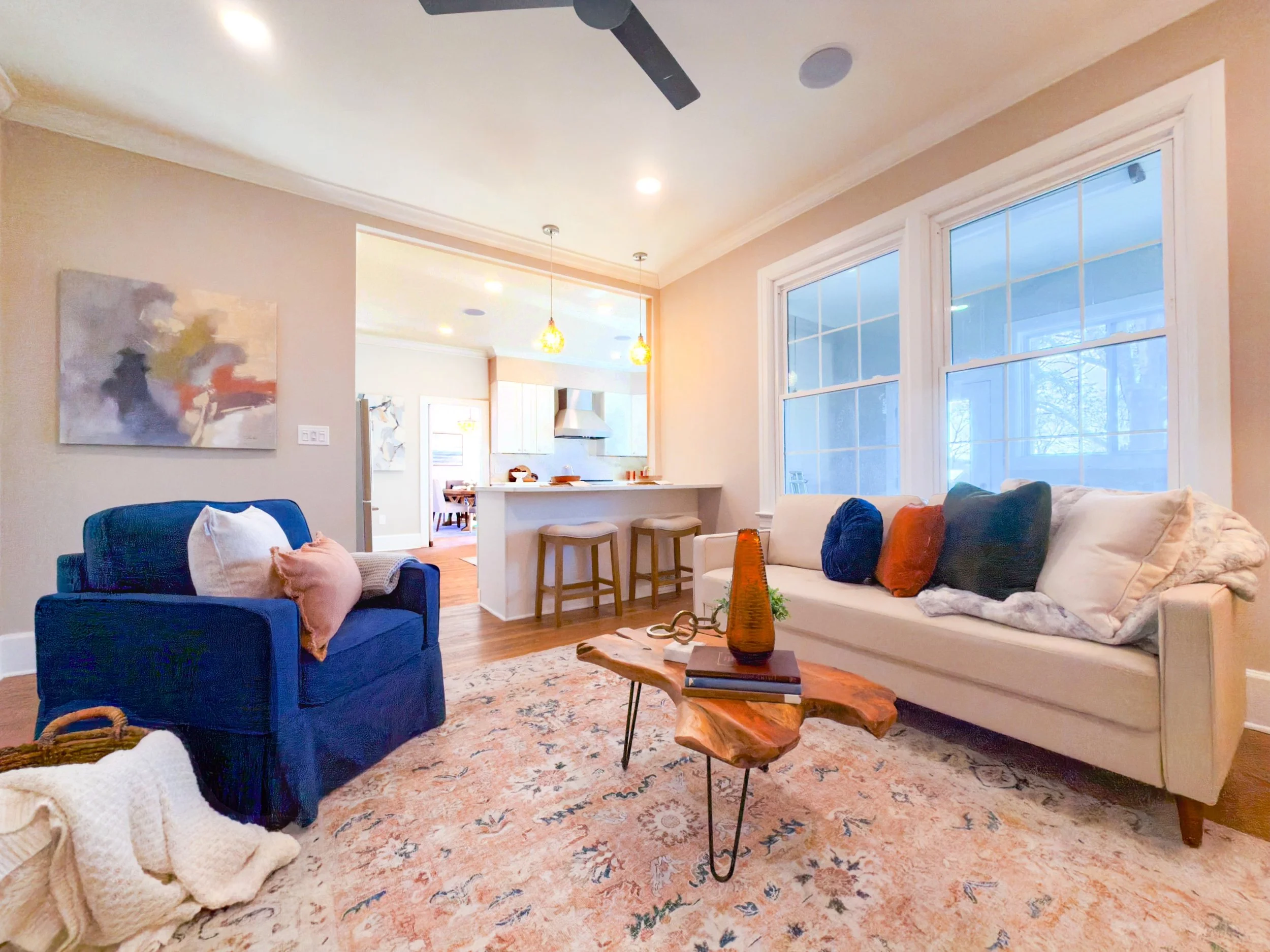 Living room with beige walls, large windows, white sofa with colorful pillows, blue armchair with pillows, wooden coffee table with decorative items, an abstract painting on the wall, and a view into the kitchen area with pendant lights.