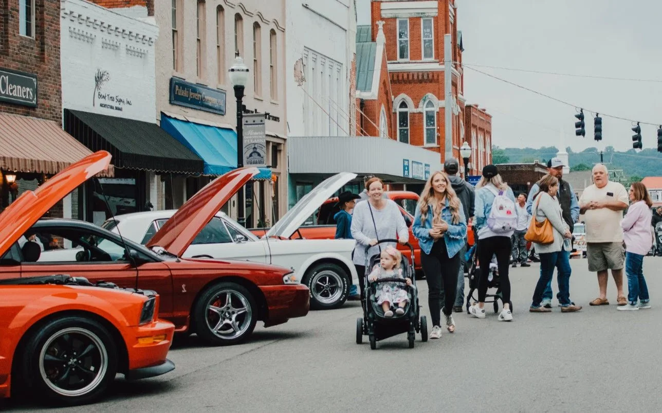 People walking along a street lined with vintage cars parked with their hoods open in a small town. Some people are chatting, and a woman is pushing a stroller with a young girl.