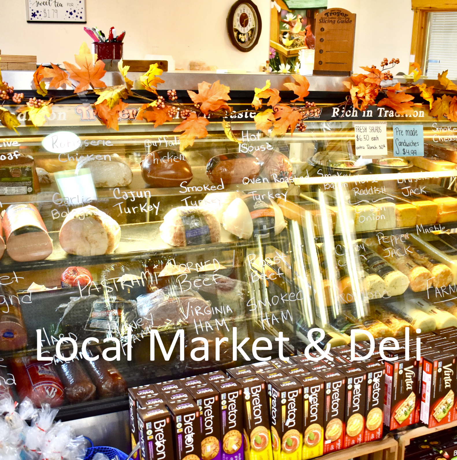 Display case at a local market or deli with a variety of meats such as chicken, turkey, ham, smoked, and roast beef, along with cheese and boxed products. The display is decorated with autumn leaves and berries, and there are small signs indicating prices and types of items, with the text 'Local Market & Deli' overlayed on the image.
