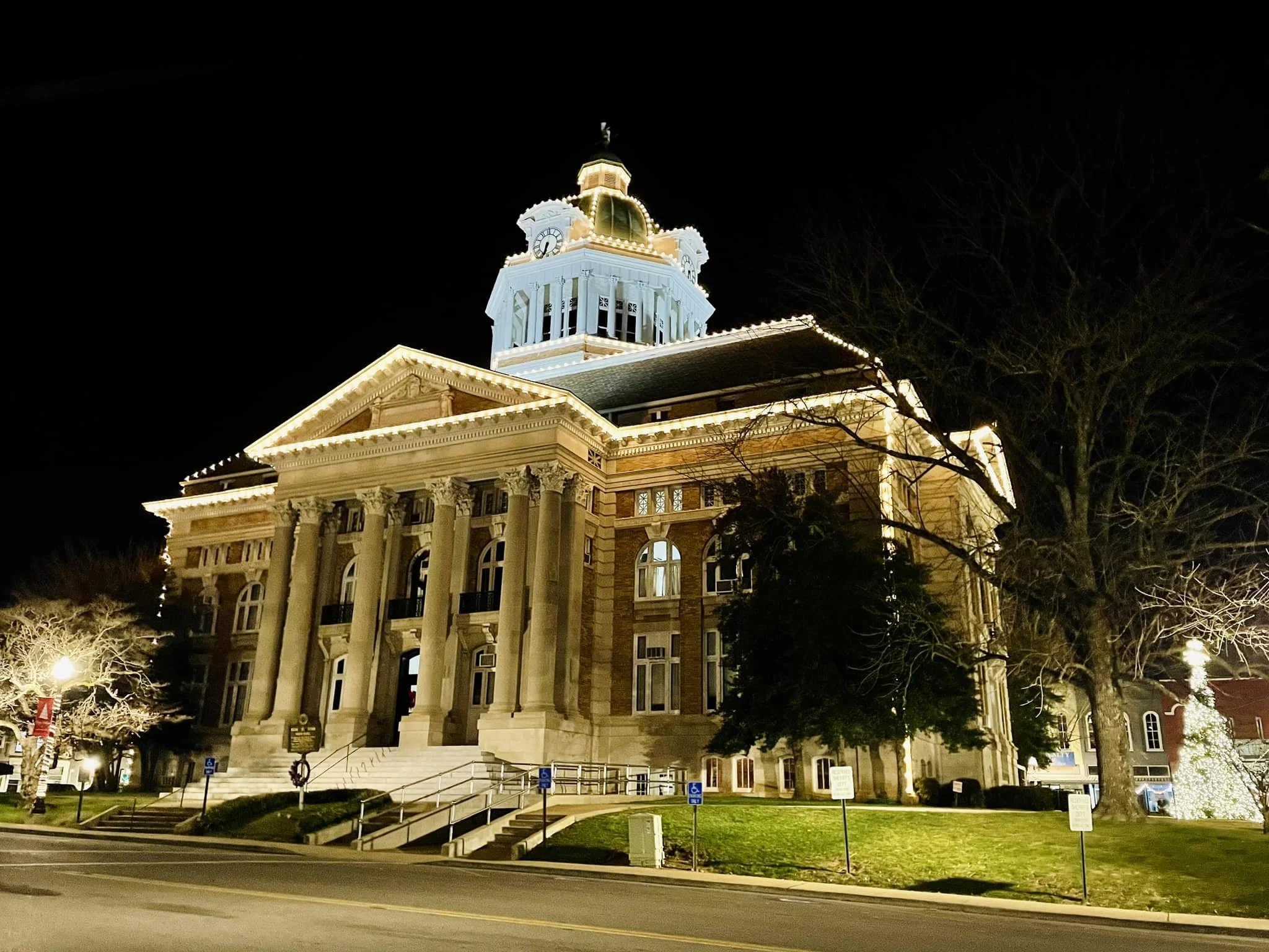 Night view of a historic courthouse building illuminated with lights, featuring tall columns and a dome on top, with a tree and holiday lights in the foreground.