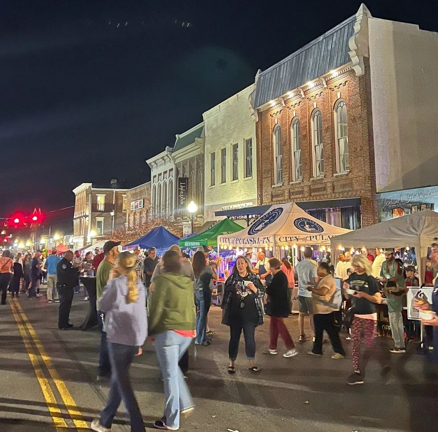 Night scene of a busy street festival with numerous vendor tents and people walking, some in conversation, across the street. Old brick and historic style buildings line the background, illuminated by streetlights.