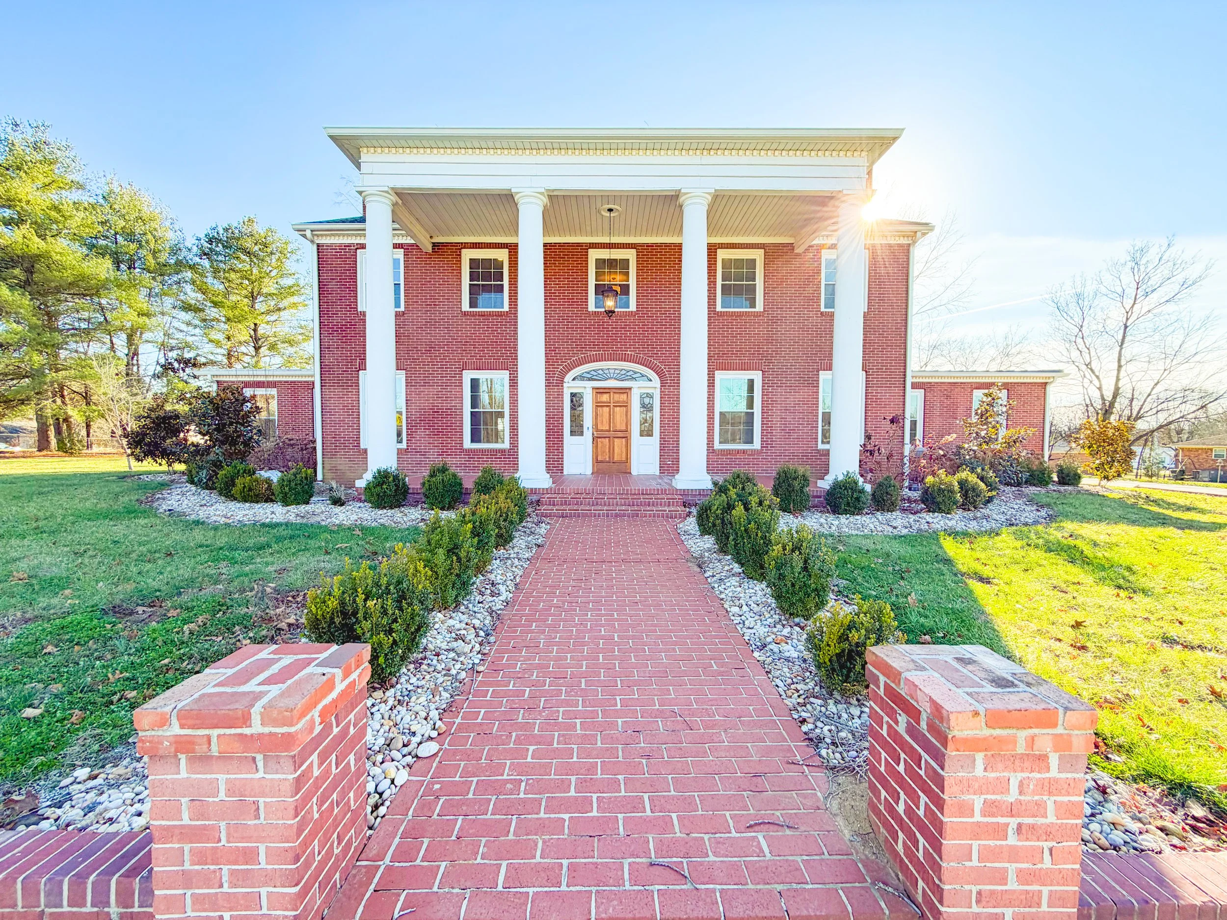 Front view of a large red brick house with white columns, a brick pathway, and landscaped yard with bushes and trees in bright sunlight.