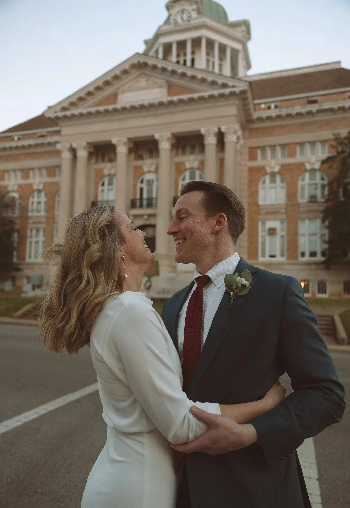 A couple in wedding attire standing outside in front of a large, historic brick building with columns and a clock tower, smiling and embracing.
