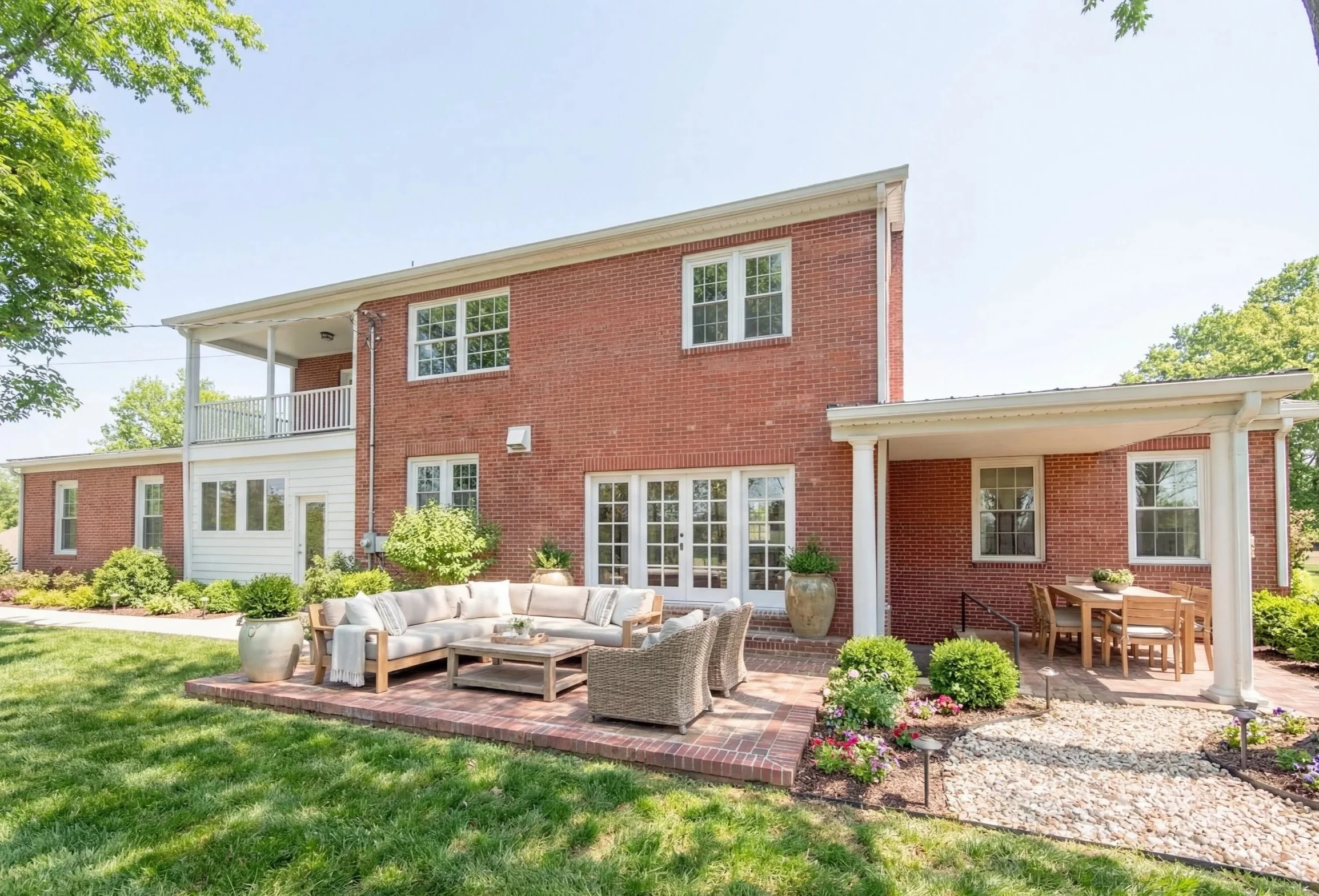 Backyard of a two-story brick house with outdoor furniture, potted plants, and landscaped garden, on a sunny day.