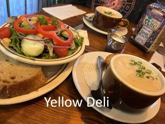 A restaurant table at Yellow Deli in Pulaski, TN with a salad, a slice of bread, and two cups of soup. One cup of soup has green onion garnish, and the other is a dark coffee. There are utensils, a napkin, a sugar container, and a menu on the table.