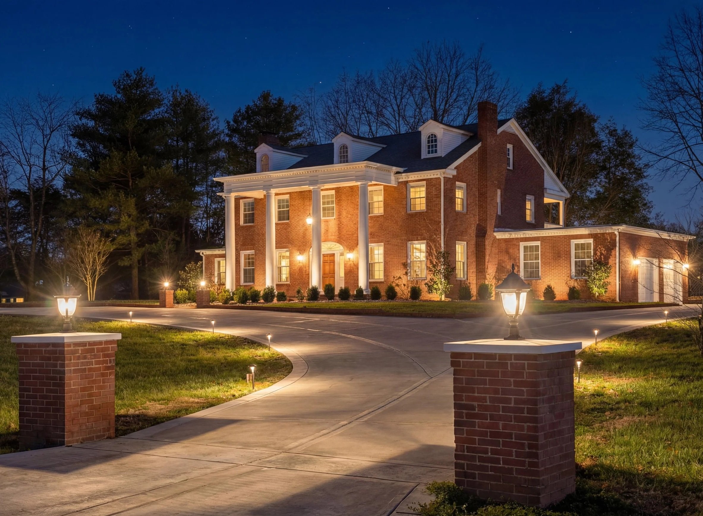 A large brick house at night illuminated by exterior lights, with a curved driveway, lamp posts, and a well-maintained lawn surrounded by trees.