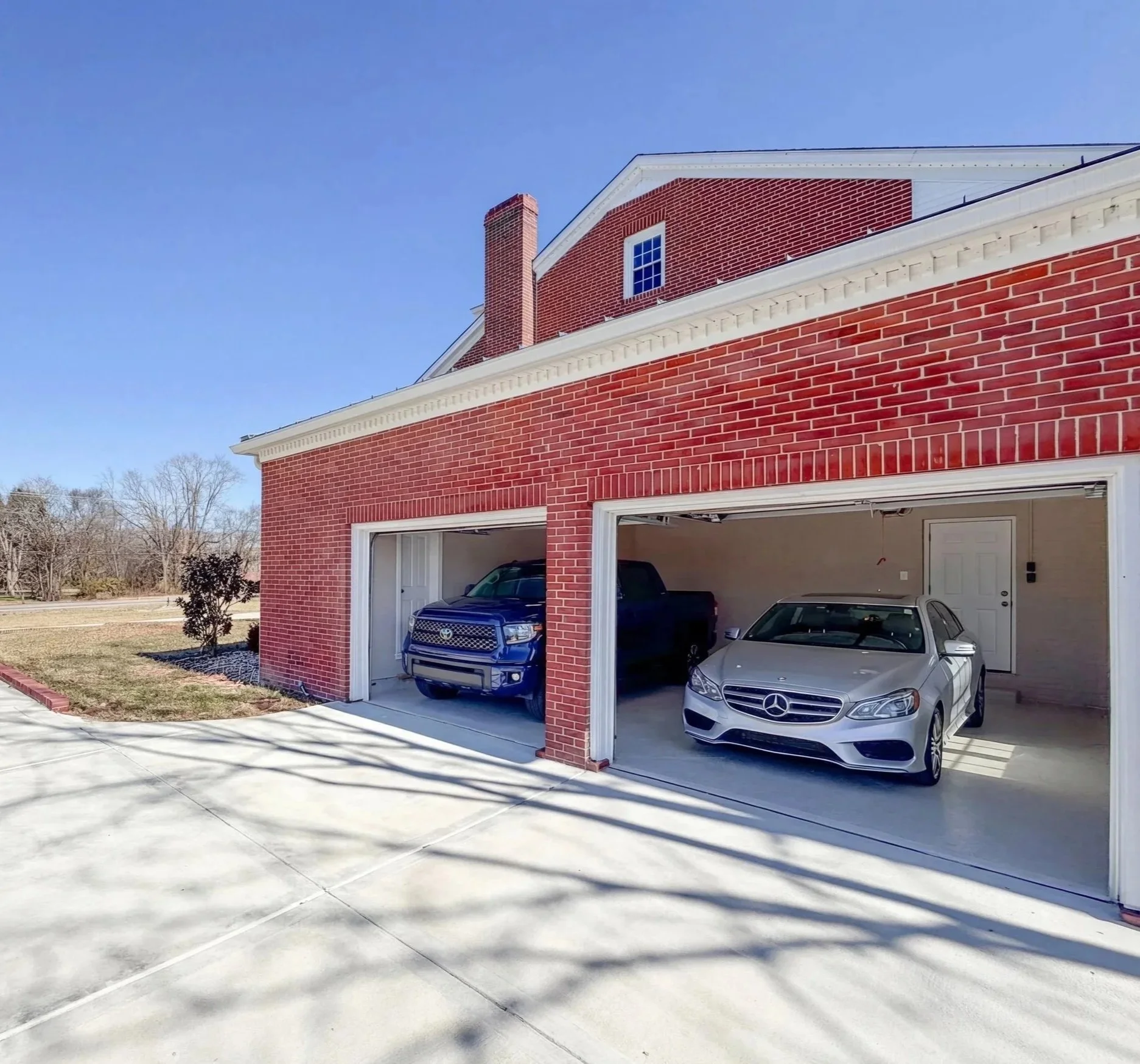 A red brick house with a double garage containing three parked cars: a blue pickup truck, a dark gray pickup, and a silver Mercedes-Benz.
