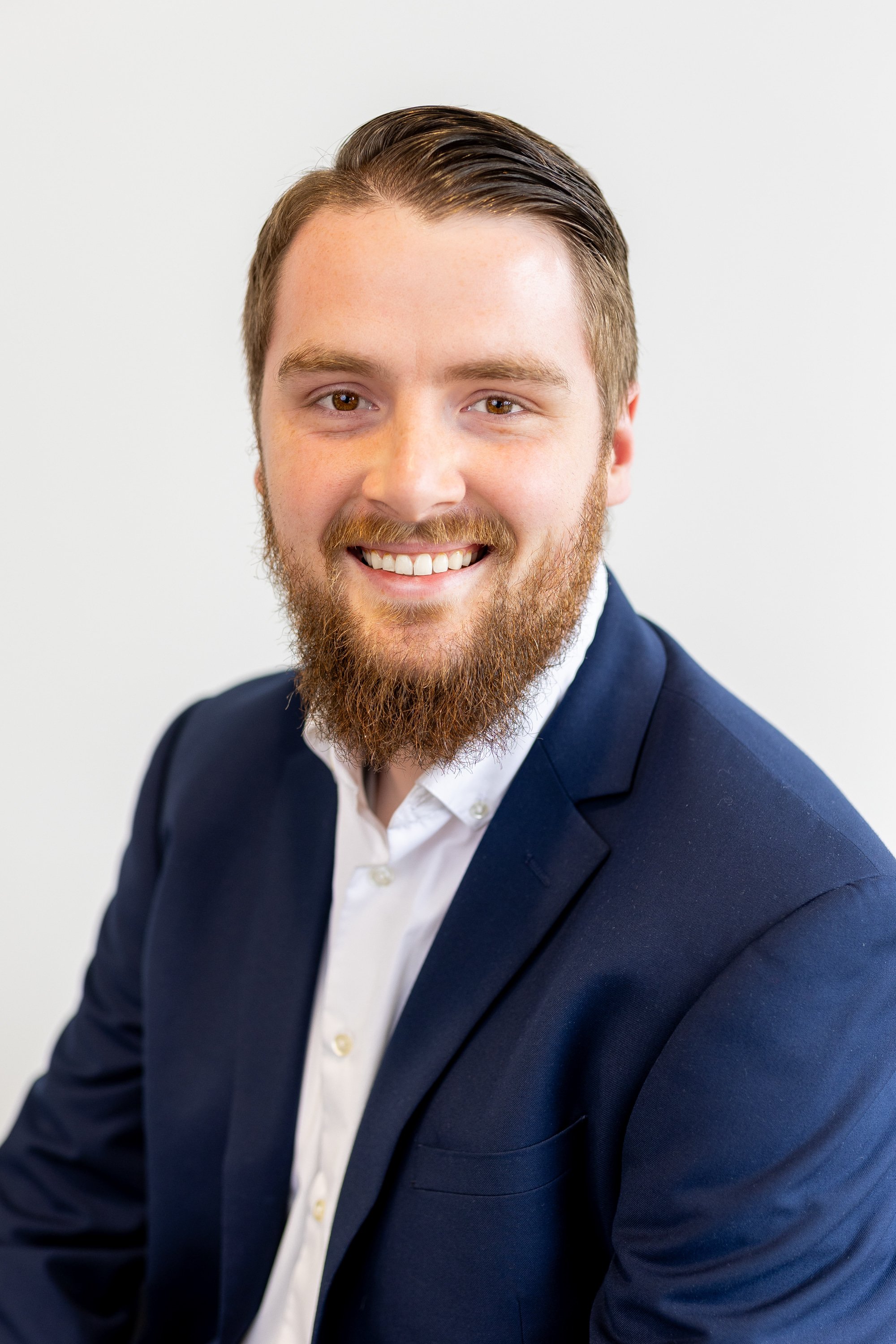 Headshot of a smiling man with a beard, wearing a navy blue suit jacket and white shirt, against a plain light background.