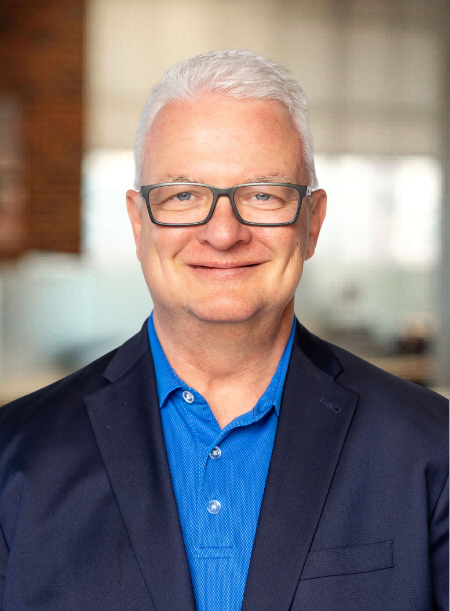 A middle-aged man with white hair and glasses, wearing a dark blazer and a blue shirt, smiling at the camera in a professional setting.