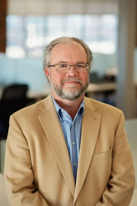 A middle-aged man with glasses, a beard, and gray hair, wearing a tan blazer over a blue shirt, standing in an office environment.