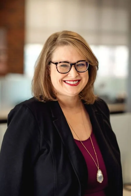 A smiling woman with shoulder-length blonde hair, glasses, and red lipstick, wearing a black blazer and a maroon top with a necklace, standing in an office environment.