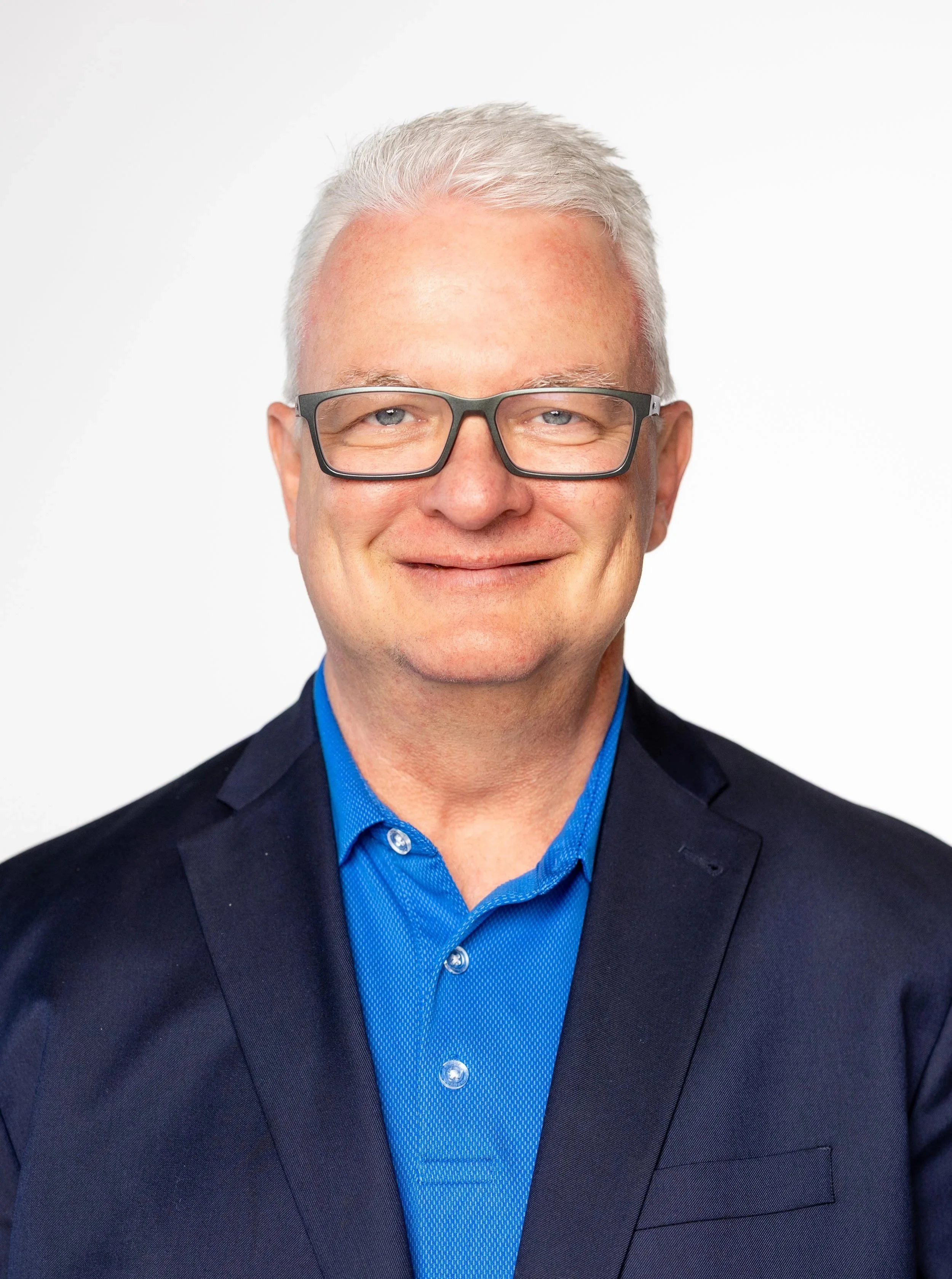 Close-up of a smiling middle-aged man with gray hair, wearing black eyeglasses, a dark suit jacket, and a bright blue shirt, against a plain white background.