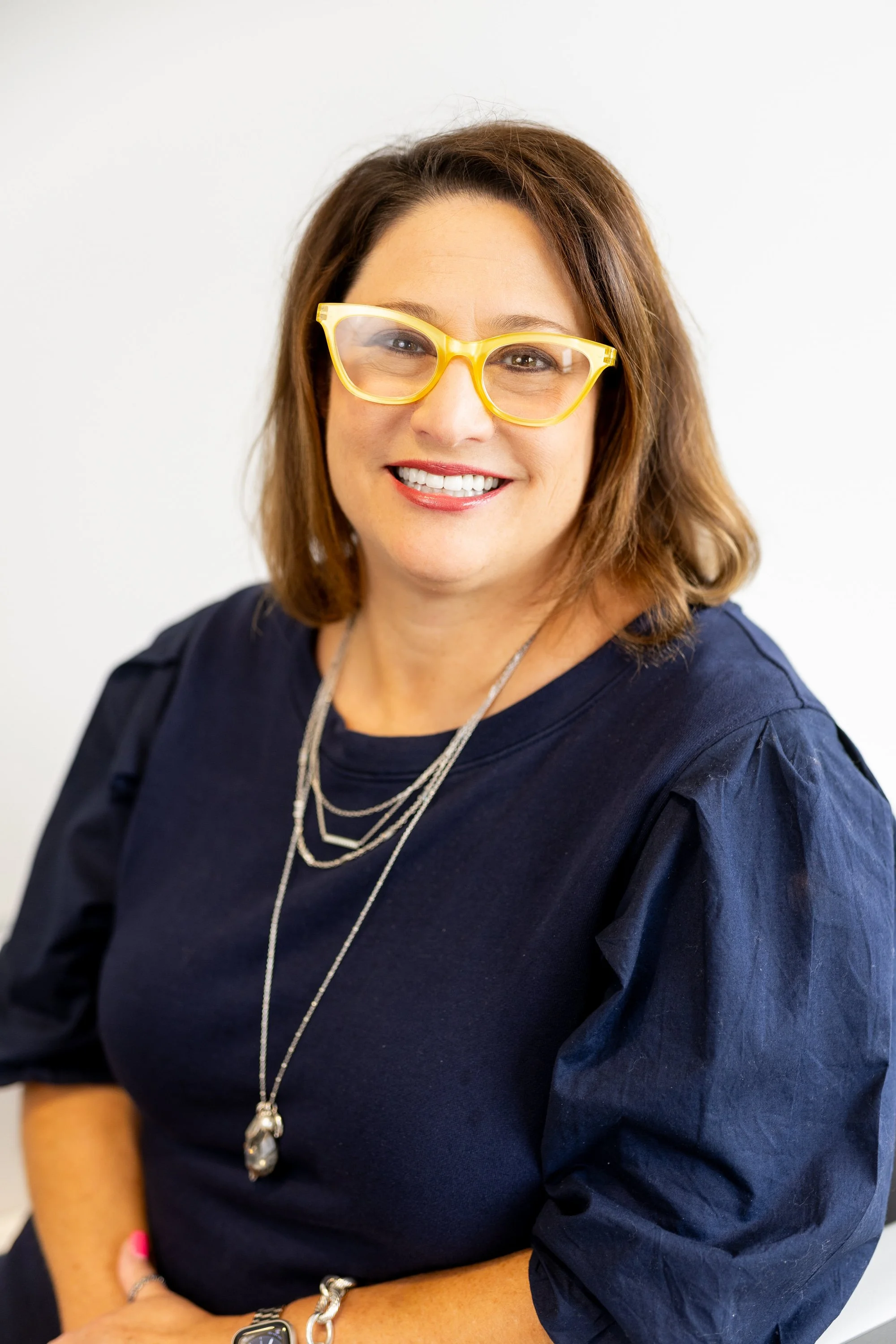 Close-up of a woman wearing yellow glasses, a dark blue top with puffed sleeves, layered necklaces, and a silver bracelet, smiling against a plain white background.