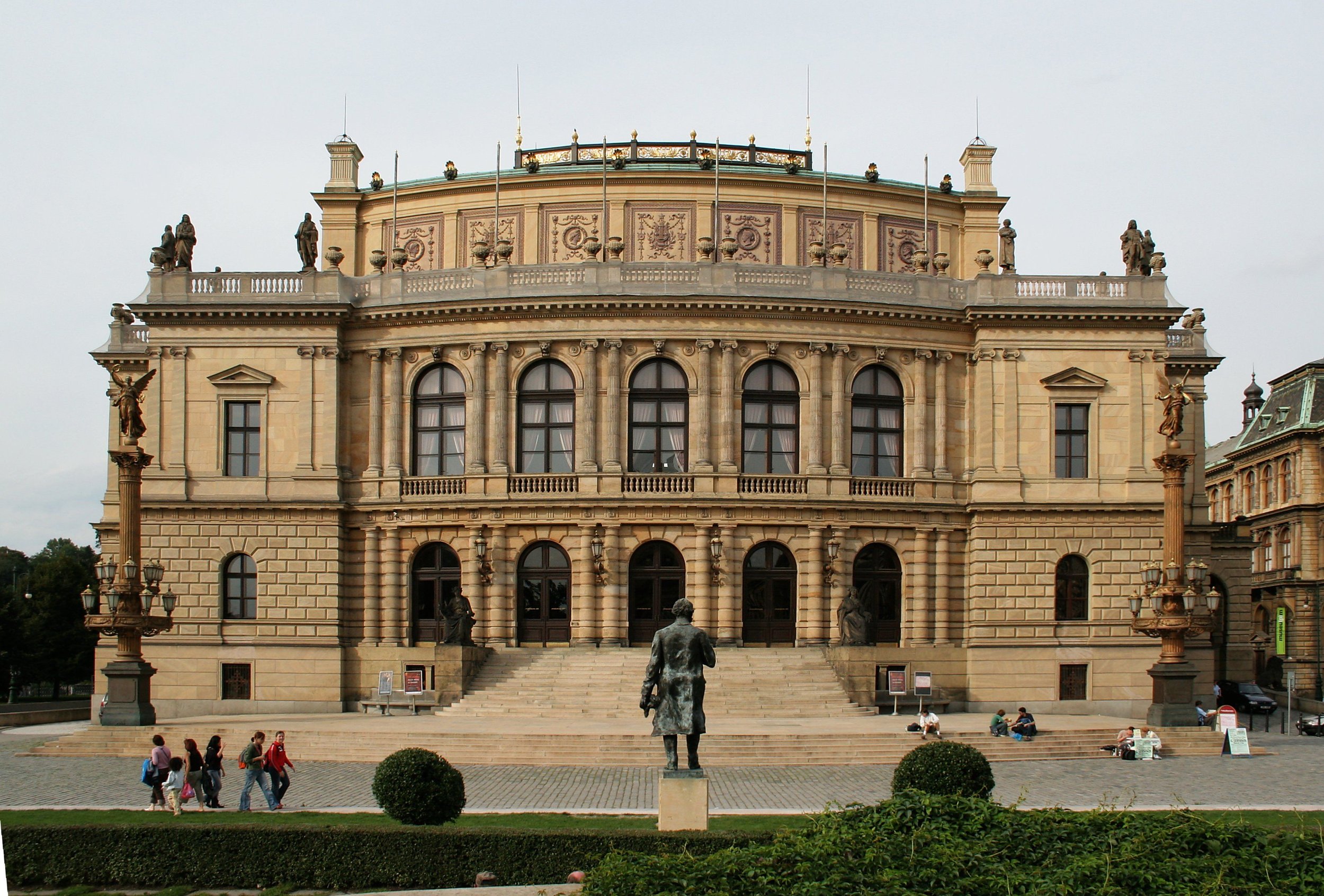 Historical building with ornate architecture and statues on the roof, entrance steps, surrounded by people sitting and walking, with greenery in the foreground.