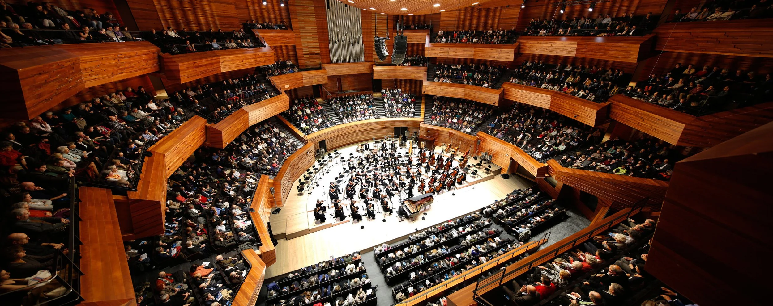 Concert hall filled with audience watching an orchestra on stage, with wood interior design.