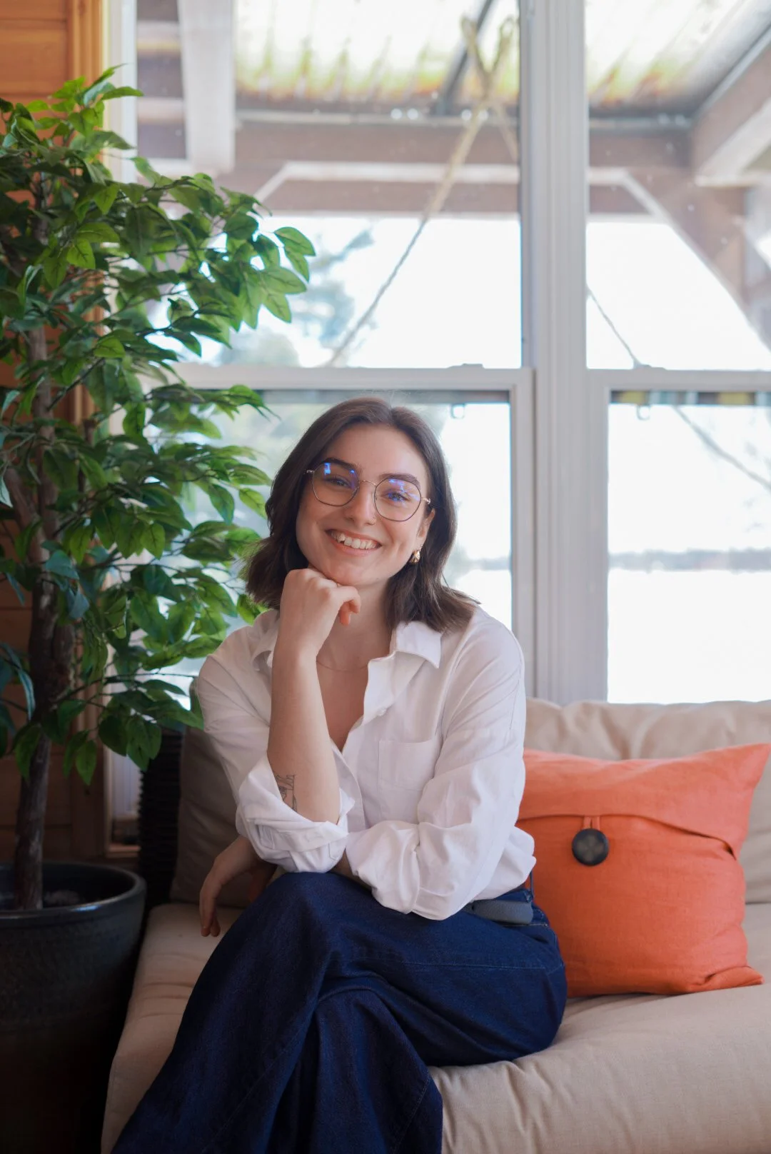 Une femme souriante, portant des lunettes et une chemise blanche, assise sur un canapé dans une pièce lumineuse avec une grande fenêtre et une plante verte à côté.