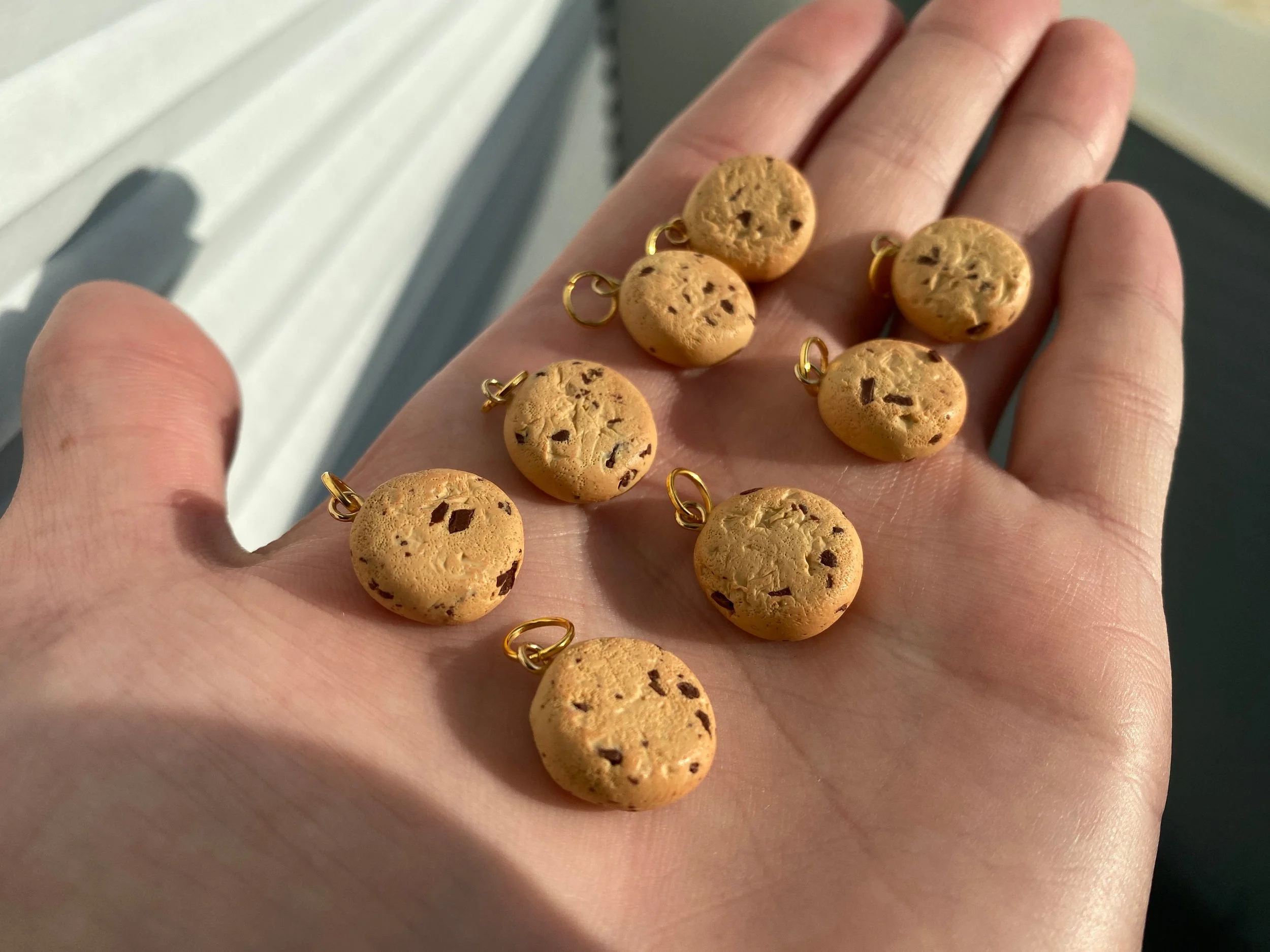 A hand holding several small, round, chocolate chip cookie charms with gold loops for jewelry, set against a background of a building with siding.