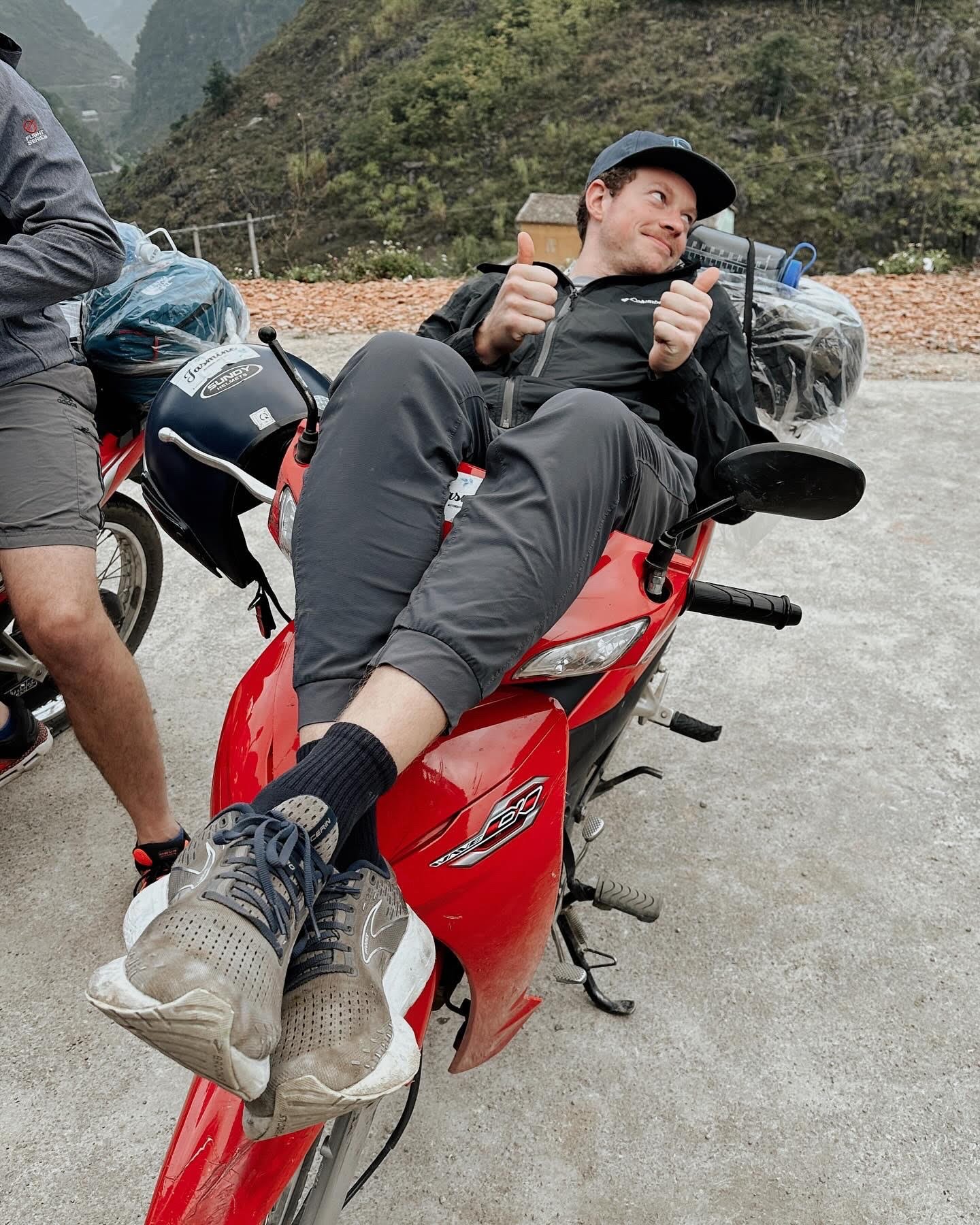 Person sitting on a red scooter giving thumbs up, with a backpack and a helmet nearby, in a mountainous outdoor setting.