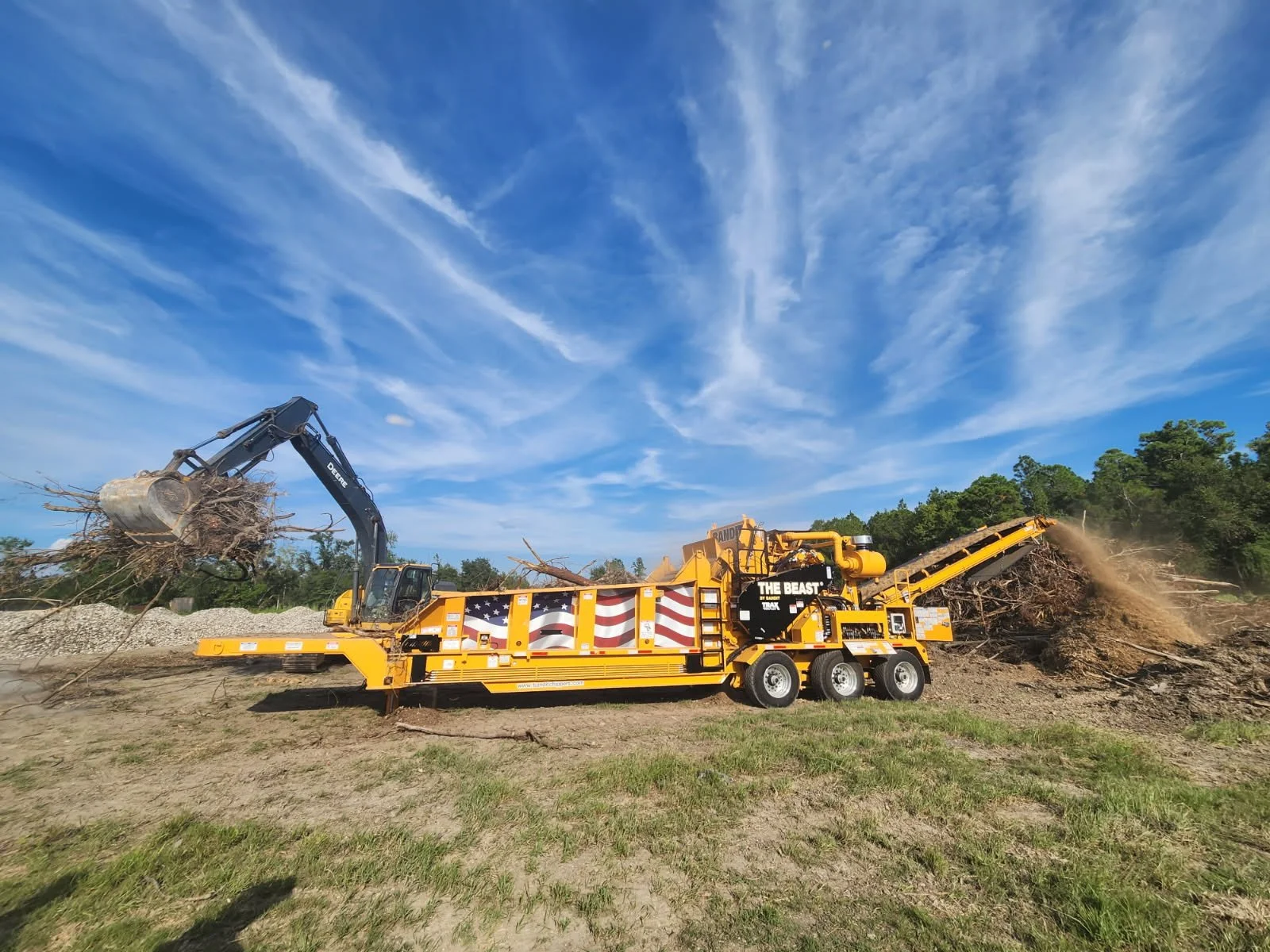 A large yellow industrial wood chipping machine with an American flag design on its side working outdoors, with a black excavator lifting tree branches into it, against a background of blue sky and green trees.