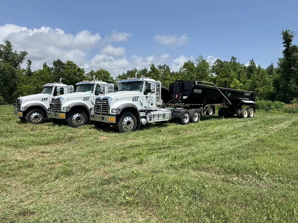 Three white Mack trucks with black dump trailers parked on a grassy field under a partly cloudy sky, surrounded by trees.