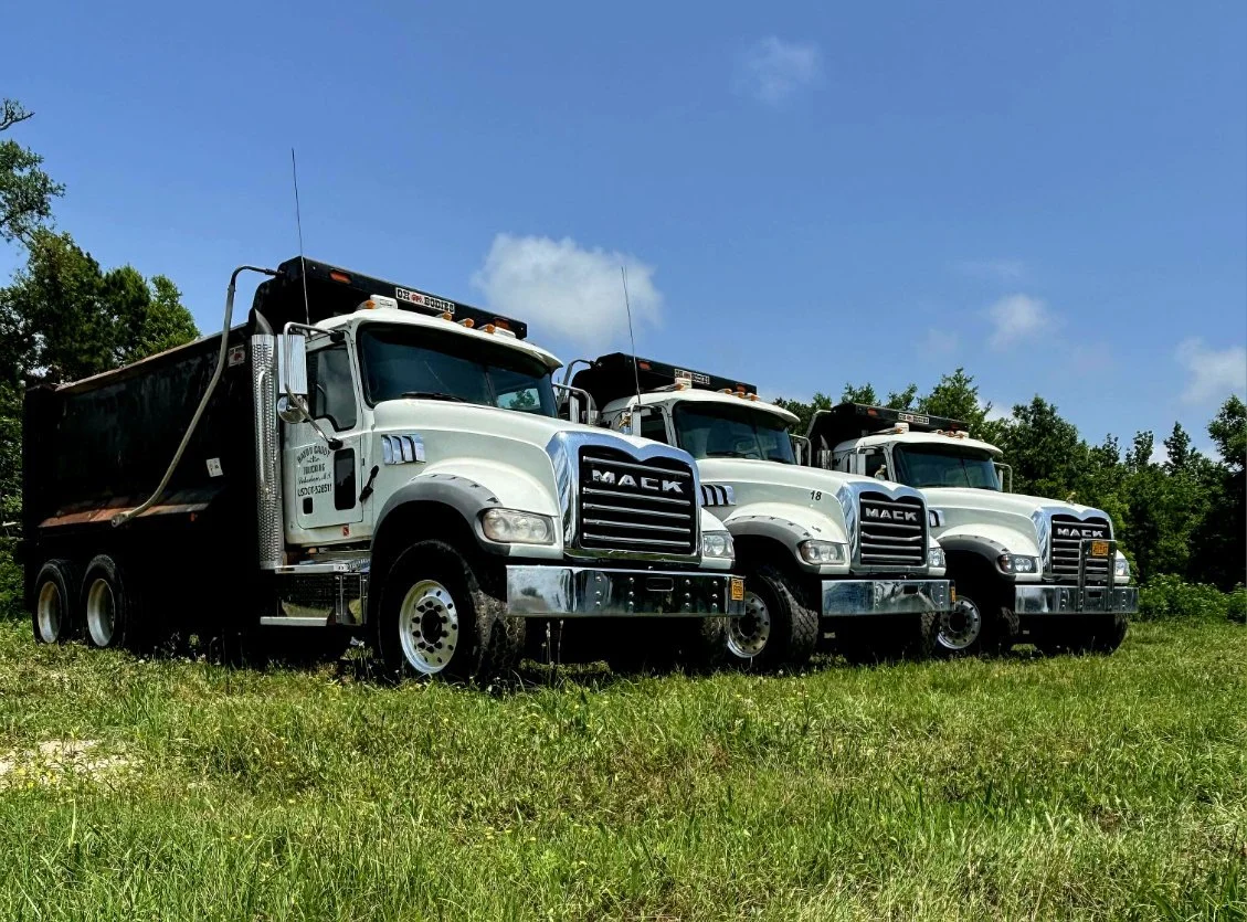 Three white Mack semi-trucks parked on a grassy field under a blue sky with trees in the background.