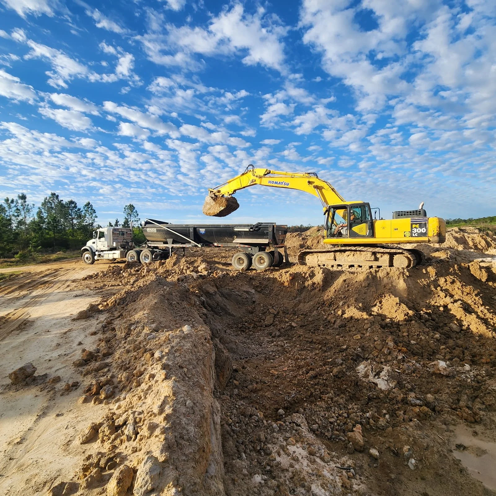 A yellow excavator digging soil at a construction site with a blue sky and scattered clouds in the background.