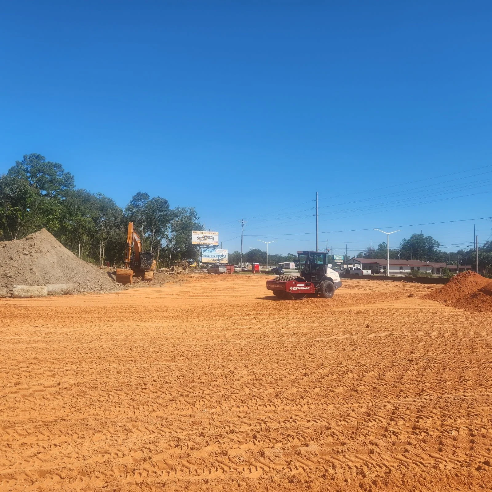 Construction site with a dirt road, construction vehicles, piles of dirt, and trees in the background under a clear blue sky.
