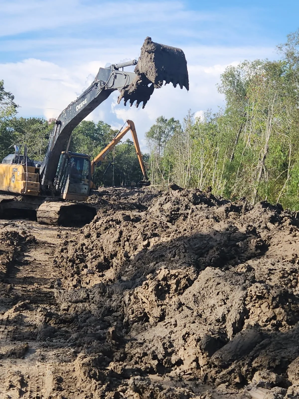 A John Deere excavator with a large bucket lifting dirt at a construction site surrounded by trees.