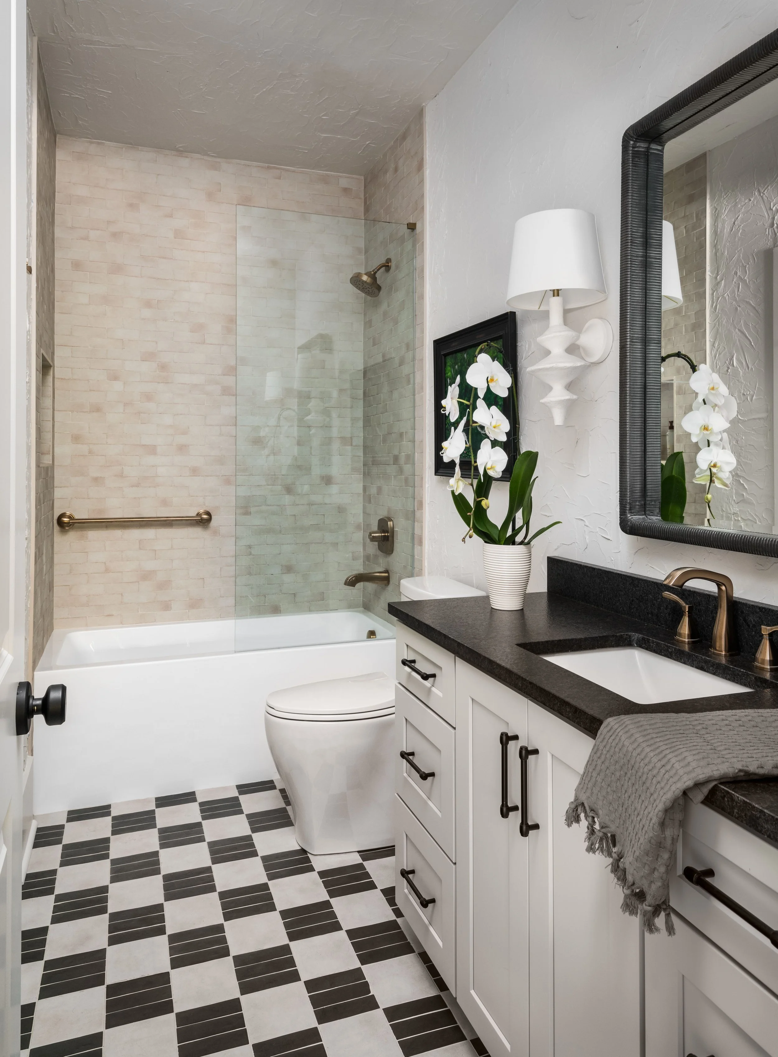 Bathroom with a glass-enclosed shower, black and white checkered floor, white vanity with black countertop, a white orchid plant, wall-mounted lamp, large mirror, and decorative framed artwork.