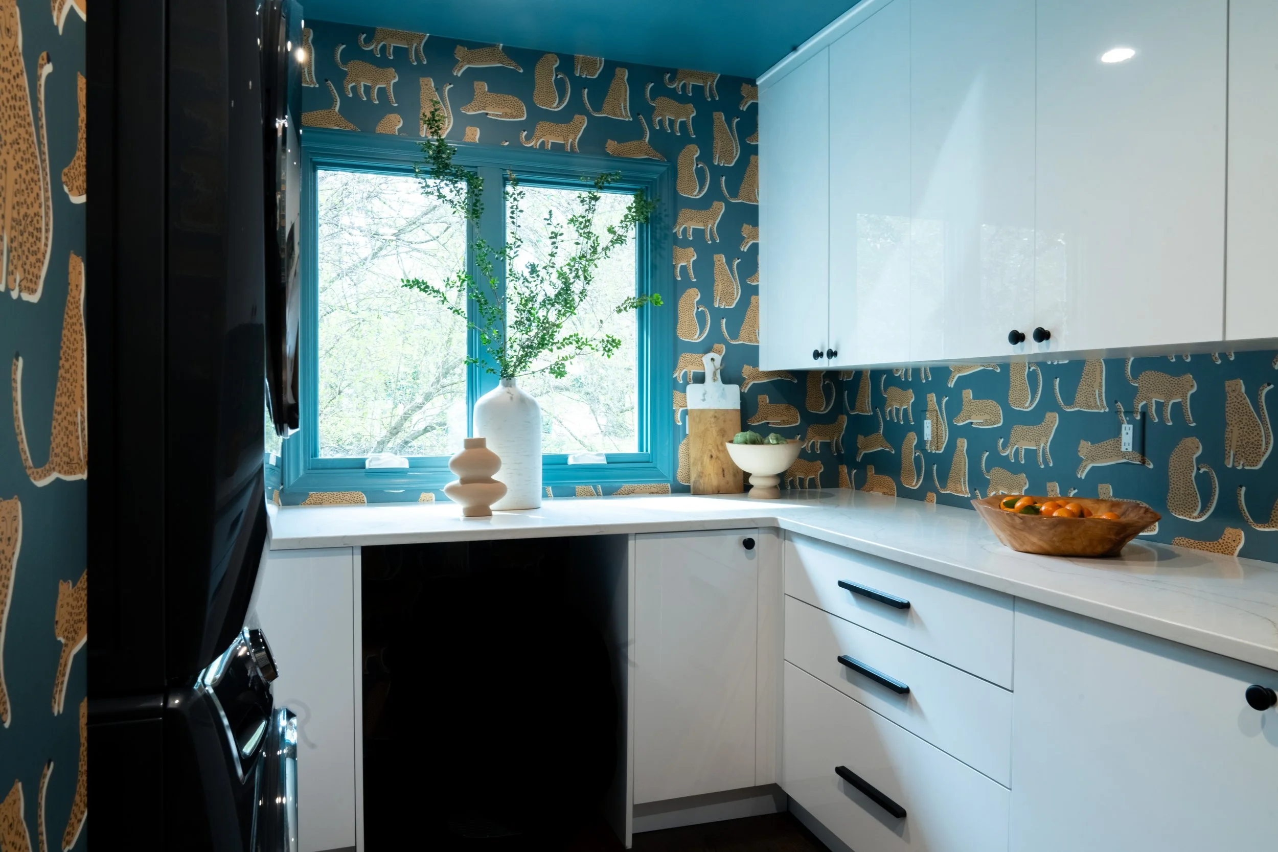A kitchen with a blue ceiling and leopard print wallpaper, white cabinets, and a window with greenery outside. The countertop has decorative vases, a wooden cutting board, and a bowl of oranges.