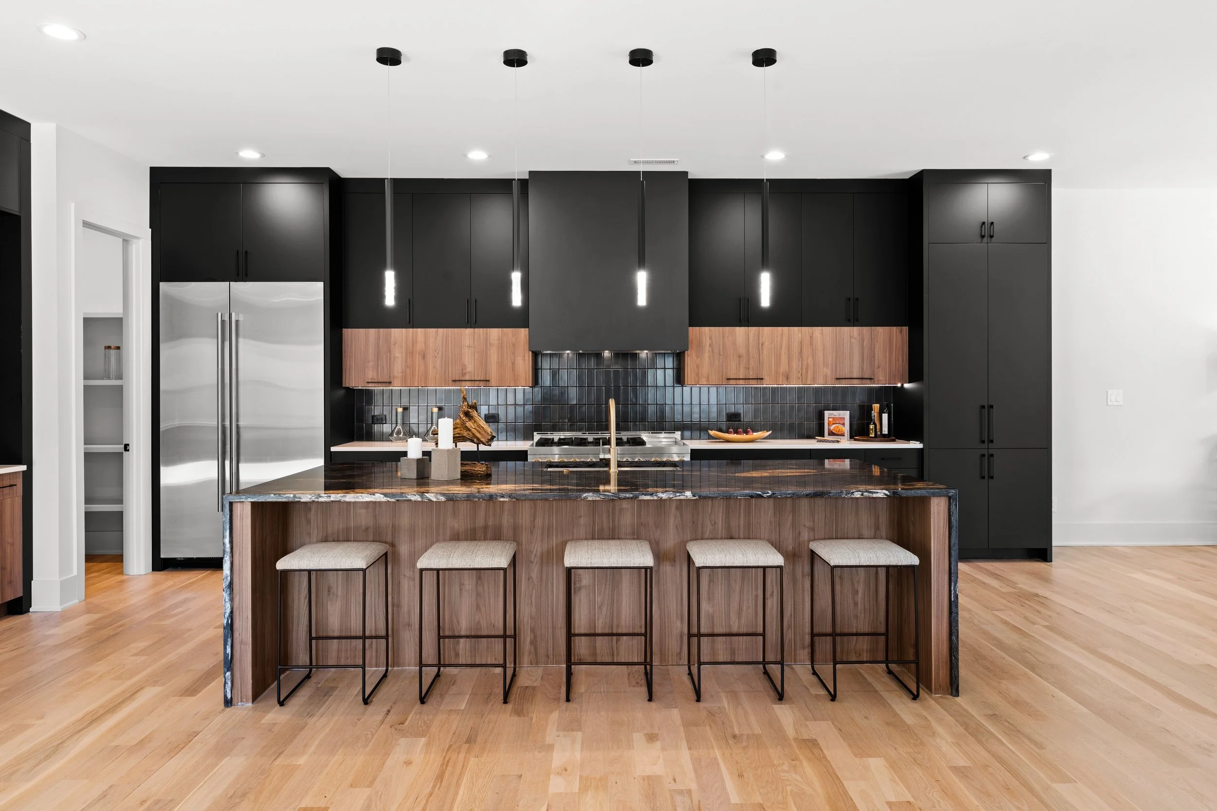 Modern kitchen with black upper and lower cabinets, a kitchen island with a dark wood finish and marble countertop, black pendant lights hanging above, and hardwood flooring.